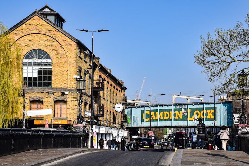 A street scene in Hampstead featuring a historic brick building with large arched and rectangular windows, some with shutters, and a small tower with a peaked roof on the left. The foreground shows a 
